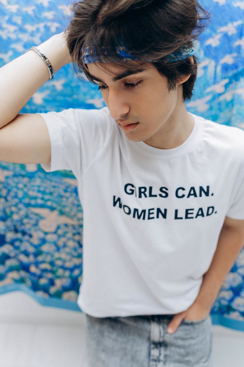 Close-up of Jamal looking down, a blue scarf in his hair, in a white T-shirt that reads “Girls can. Women lead.”