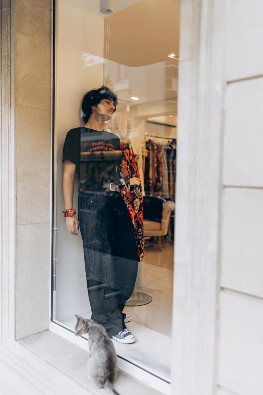 Jamal stands inside the shop window in a black slogan T-shirt while a grey cat sits on the step outside.