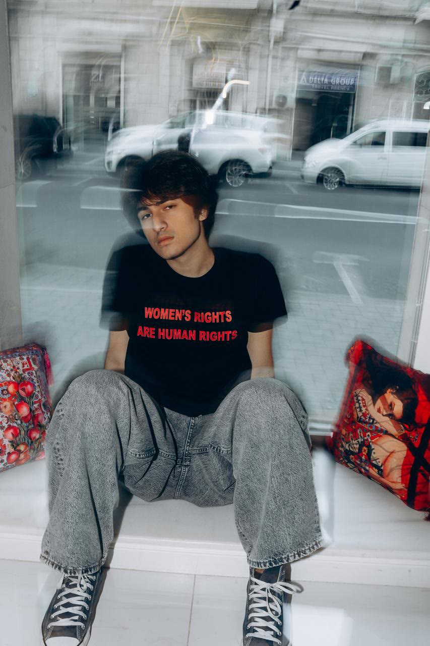 Jamal sits on the windowsill between two printed cushions in a black T-shirt that reads “Women's rights are human rights”.